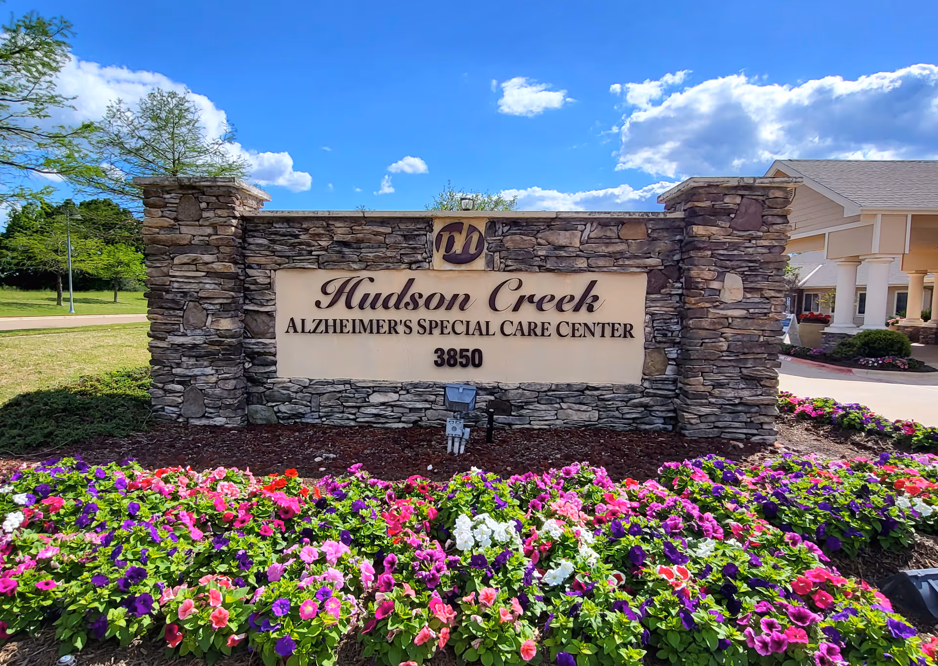 Stone sign for Hudson Creek Alzheimer's Special Care Center with the address 3850, surrounded by colorful flowers and greenery under a blue sky with clouds. Part of the building entrance is visible on the right side.