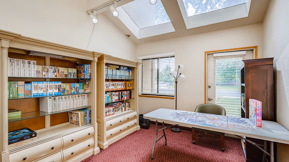 A bright room with two large skylights and a window with blinds. The room features two beige bookshelves filled with numerous puzzle boxes. A folding table with an incomplete jigsaw puzzle and a green chair are positioned near a door with blinds. A floor lamp with three lights stands near the window.