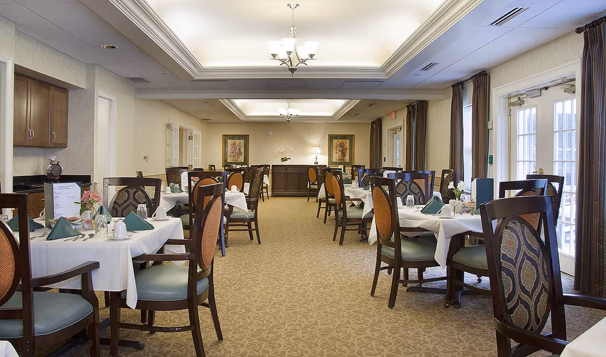 A well-lit dining room with multiple tables covered in white tablecloths, each set with green napkins, glassware, and silverware. The room features patterned carpet, wooden chairs with upholstered backs and seats, large windows with dark curtains, and framed artwork on the far wall. Ceiling lights provide soft illumination.