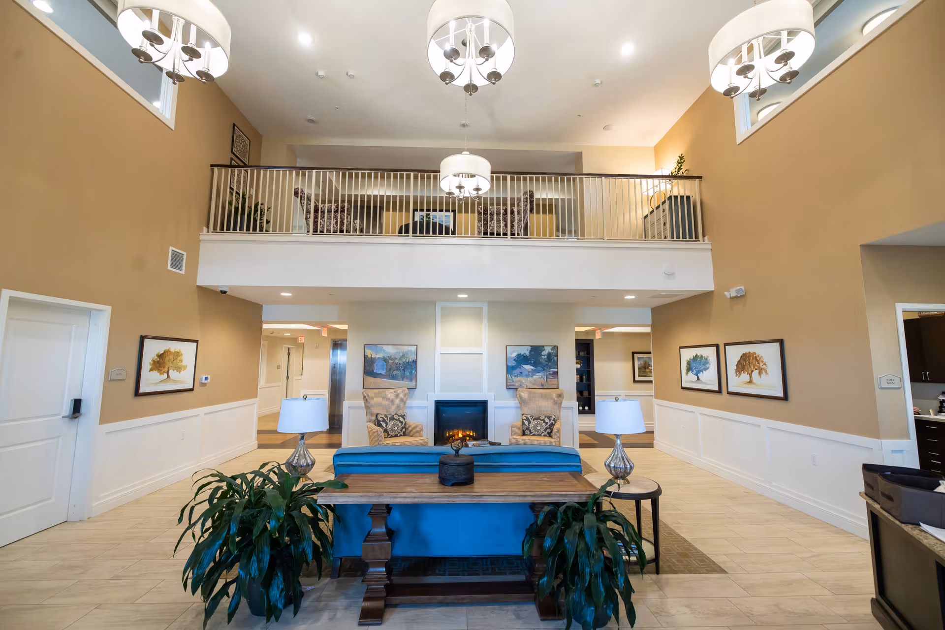 A spacious senior living facility common area with high ceilings and a mezzanine level. The room features a blue sofa, two beige armchairs with patterned cushions, a wooden table with two lamps, and two large green plants. There is a fireplace in the center of the back wall, flanked by two paintings. The walls are painted beige with white wainscoting, and there are three modern chandeliers hanging from the ceiling.