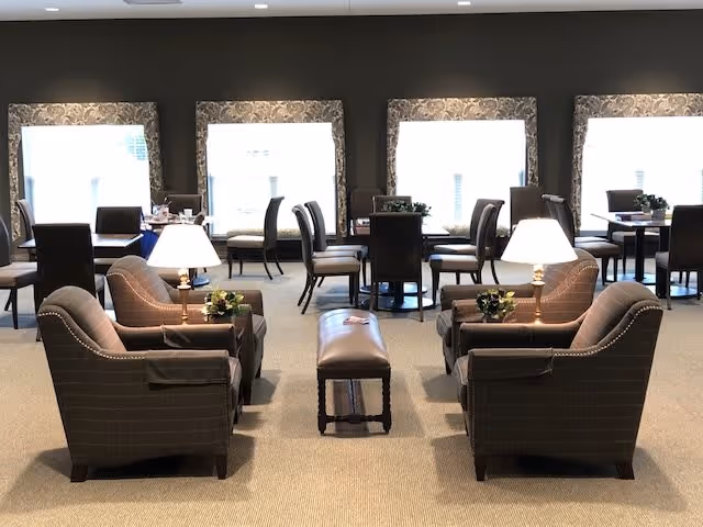 A well-lit interior room with multiple seating areas including cushioned armchairs, small tables with lamps, and dining tables with chairs near large windows covered with sheer curtains.