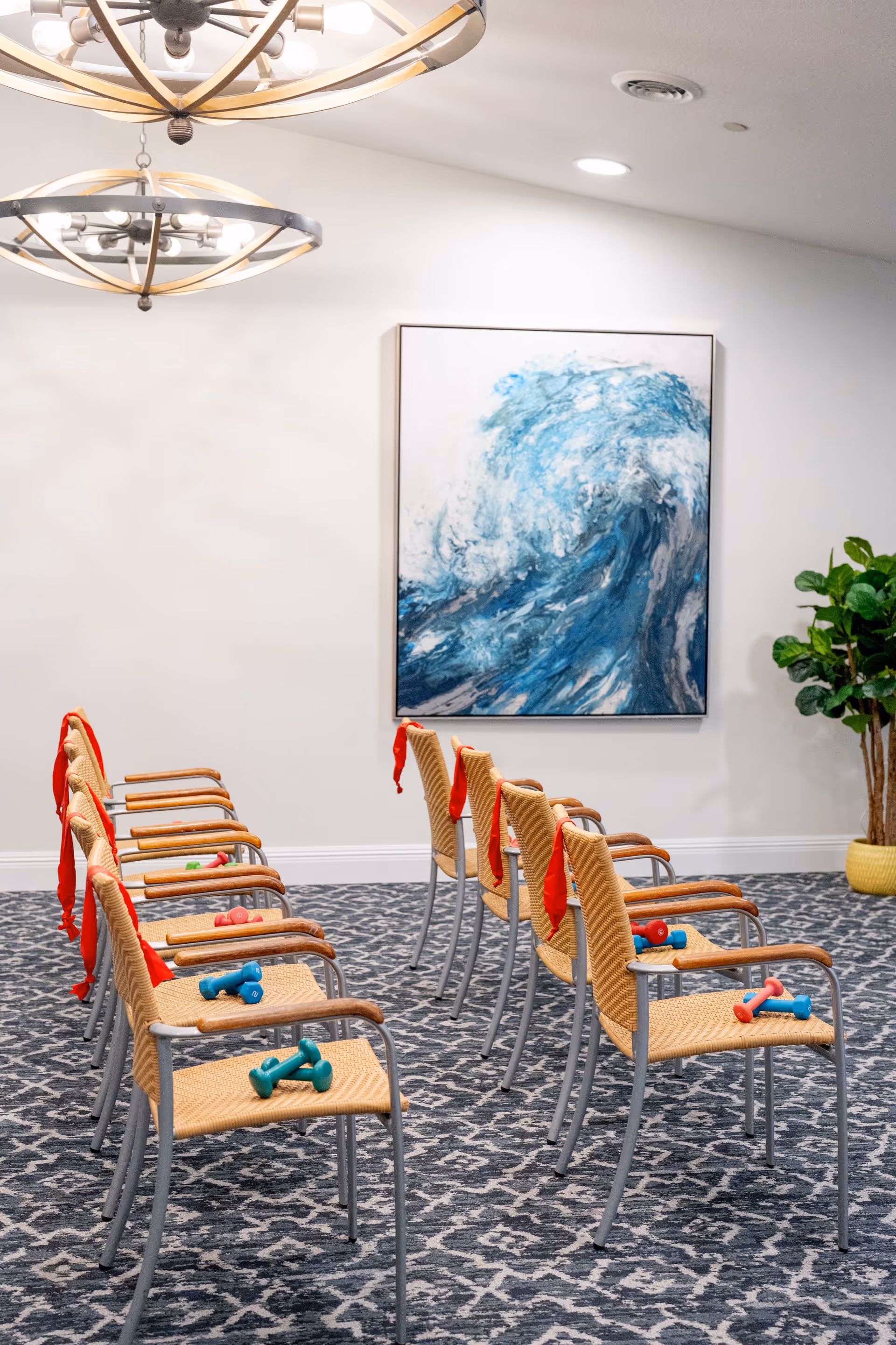 A room with two rows of wicker chairs with metal legs, each chair has a pair of small dumbbells on the seat and a red resistance band hanging on the back. The room has patterned carpet flooring, a large abstract painting of a blue wave on the wall, two modern circular chandeliers hanging from the ceiling, and a potted plant in the corner.