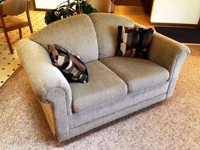 A beige upholstered loveseat with two patterned throw pillows placed on a carpeted floor in a room with a dining area visible in the background.