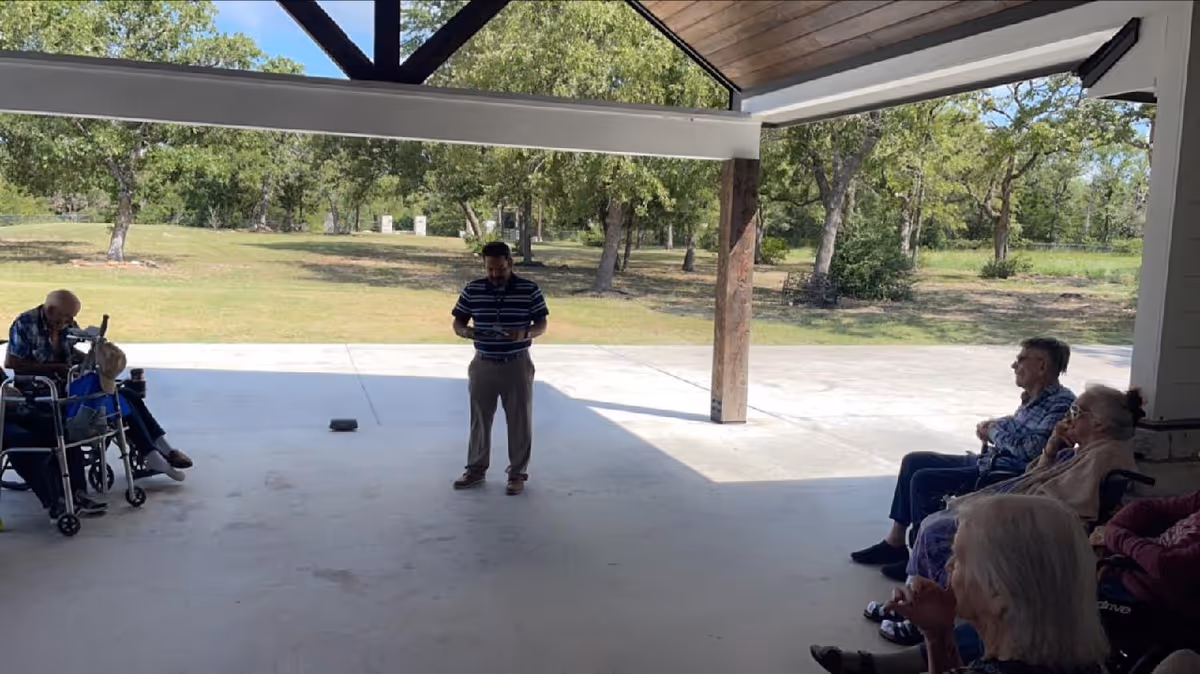 A group of elderly people sitting under a covered outdoor patio area, with a man standing in the middle holding a tablet or book. The background shows a grassy area with trees and a clear sky.