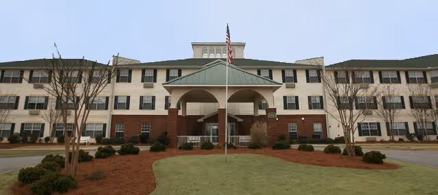 Front exterior view of The Legacy of Anderson senior living facility, a three-story building with a green roof, brick and white siding, an American flag on a flagpole in front, and landscaped grounds with small trees and bushes.