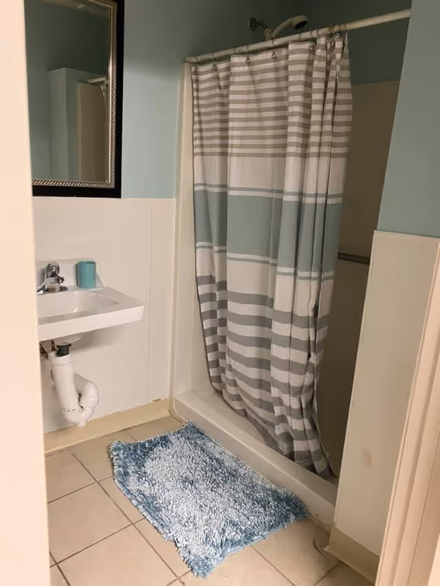A small bathroom with a white sink and faucet on the left, a rectangular mirror above the sink, a shower with a striped shower curtain in shades of gray, white, and light blue on the right, and a blue textured bath mat on the tiled floor.