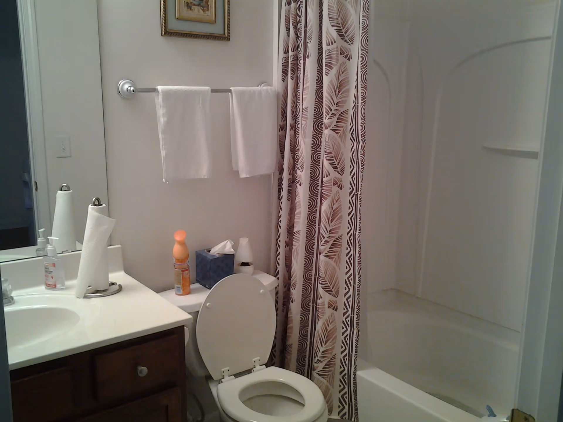A bathroom with a white toilet with the lid open, a bathtub with a patterned shower curtain, a countertop with a sink, paper towels, and hand soap. Two white towels hang on a towel rack above the toilet. There is a framed picture on the wall above the towels.