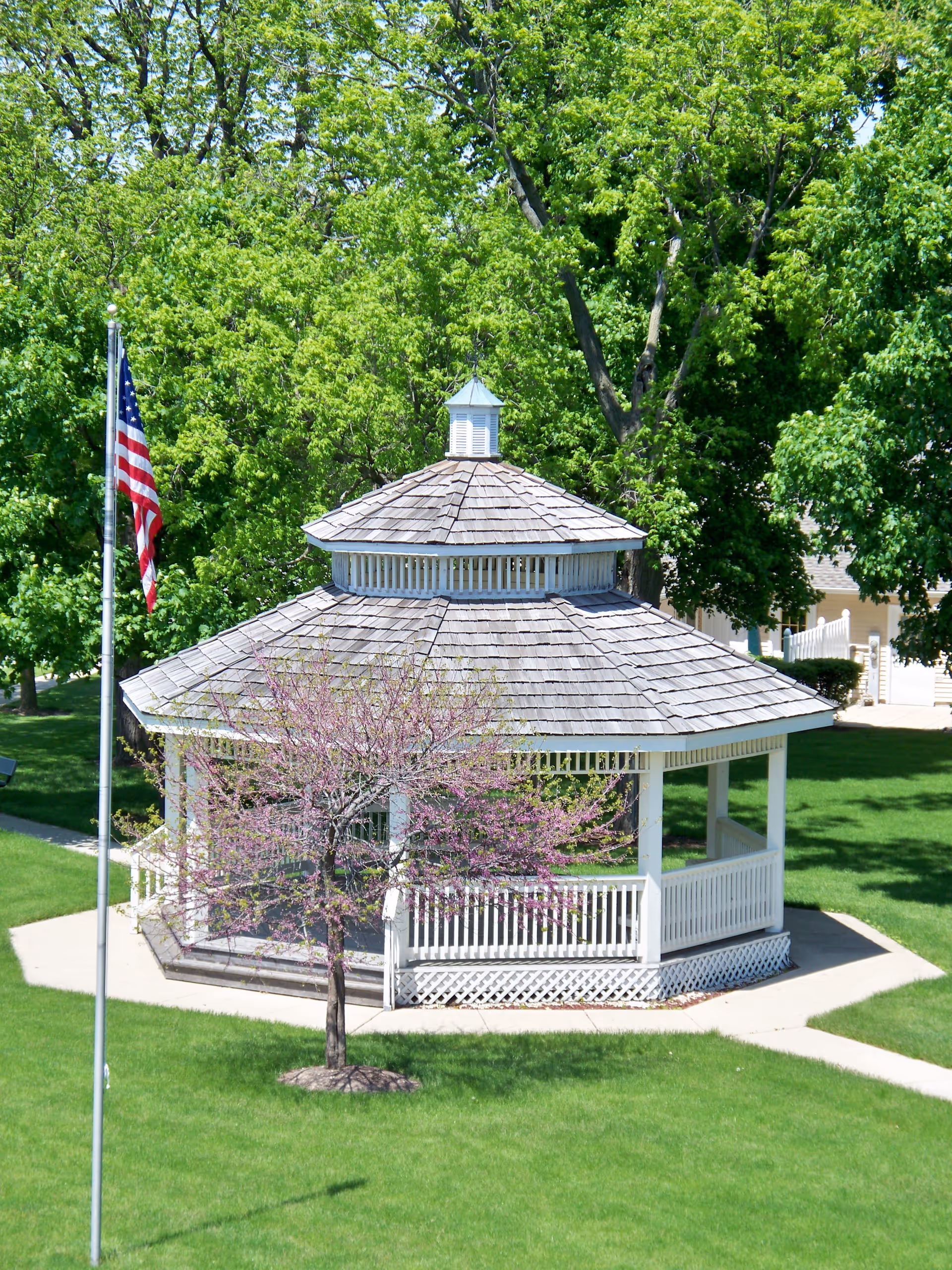 A white wooden gazebo with a shingled roof surrounded by green grass and trees. A small tree with purple blossoms is in front of the gazebo, and an American flag on a flagpole is visible to the left. The scene is bright and sunny.