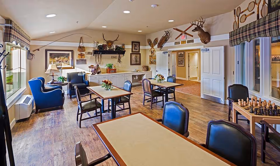 A cozy common area in a memory care community featuring several tables and chairs arranged for social activities. The room has wood flooring, large windows with plaid valances, and walls decorated with mounted deer heads, vintage snowshoes, and framed pictures. A chessboard is set up on one table, and there are comfortable armchairs near the windows. The space is well-lit with recessed ceiling lights and natural light from the windows.