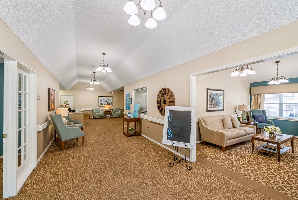Interior view of a senior living facility hallway with beige walls and carpeted floor. The hallway is furnished with several armchairs and small tables with lamps. To the right, there is an open room with a beige sofa, armchair, coffee table with flowers, and large window with curtains. A chalkboard sign with the word 'Welcome' is placed near the entrance to the room. The ceiling has multiple light fixtures with white shades.