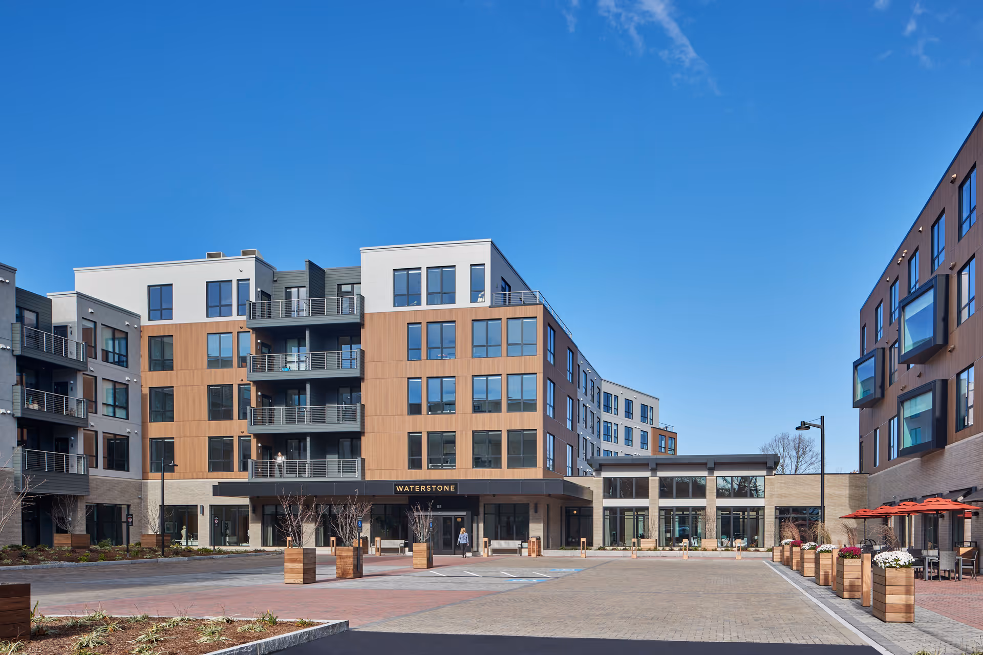 Exterior view of Waterstone of Lexington, a modern multi-story senior living facility with large windows, balconies, and a spacious paved entrance area with planters and outdoor seating under red umbrellas, set against a clear blue sky.