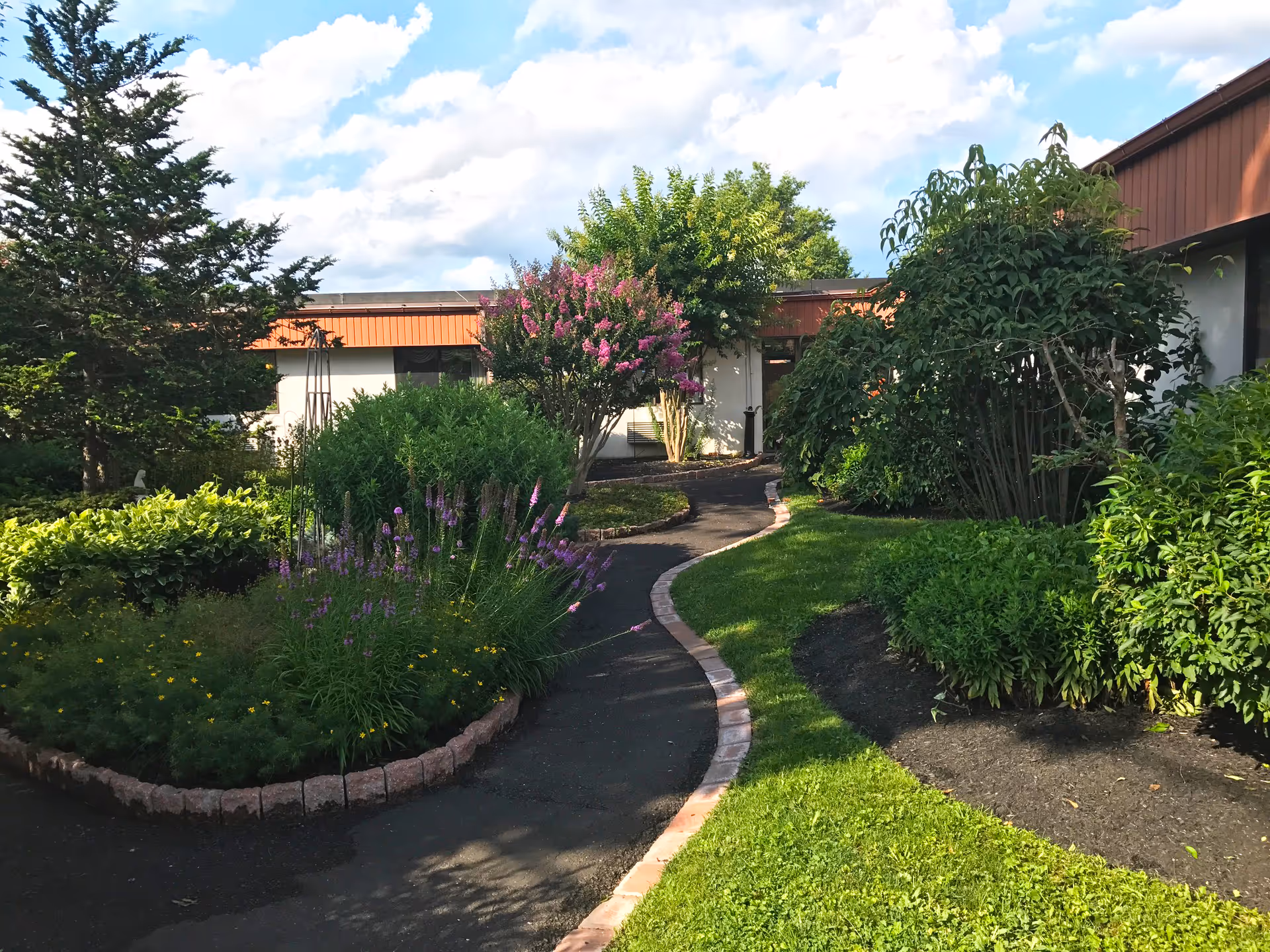 A winding paved pathway bordered by brick edging runs through a well-maintained garden with various green shrubs, flowering plants, and trees. The pathway leads to a single-story building with a brown roof under a partly cloudy blue sky.