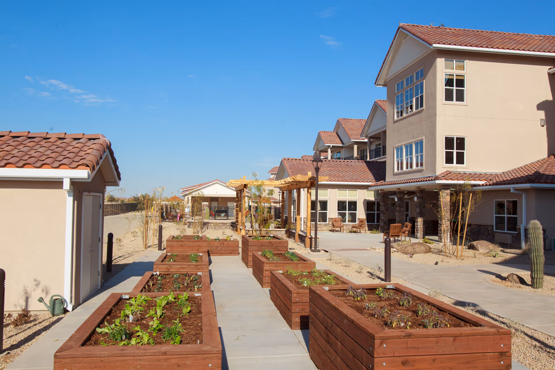 Outdoor garden area at Estrella Estates Gracious Retirement Living featuring multiple raised wooden garden beds with plants, a paved walkway, a pergola, and a multi-story building with beige walls and red tiled roofs under a clear blue sky.