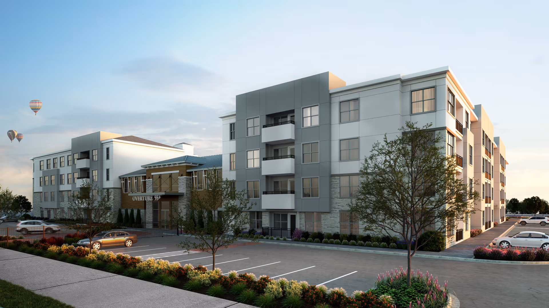 Exterior view of a modern four-story senior living facility named Overture 55+ with a parking lot in front, landscaped bushes and trees, and hot air balloons in the sky during early evening.