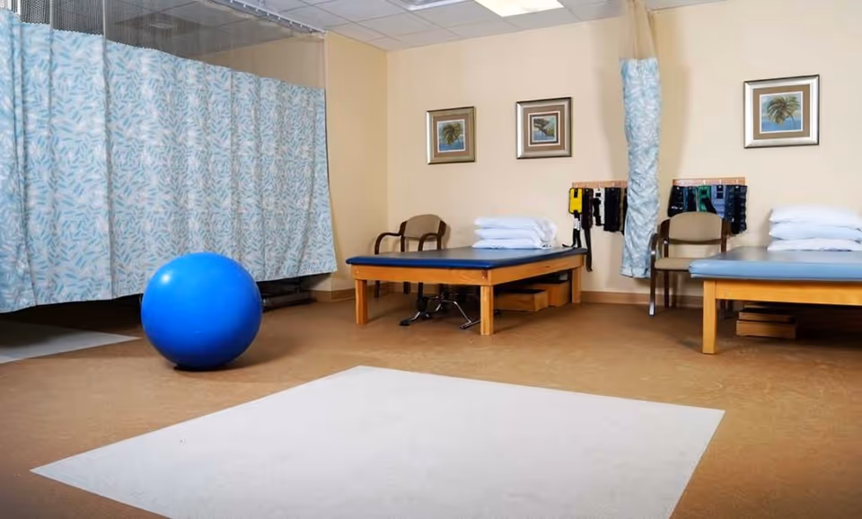 Interior therapy/exercise room with two treatment tables, stacked pillows, chairs, a large blue exercise ball, and privacy curtains.