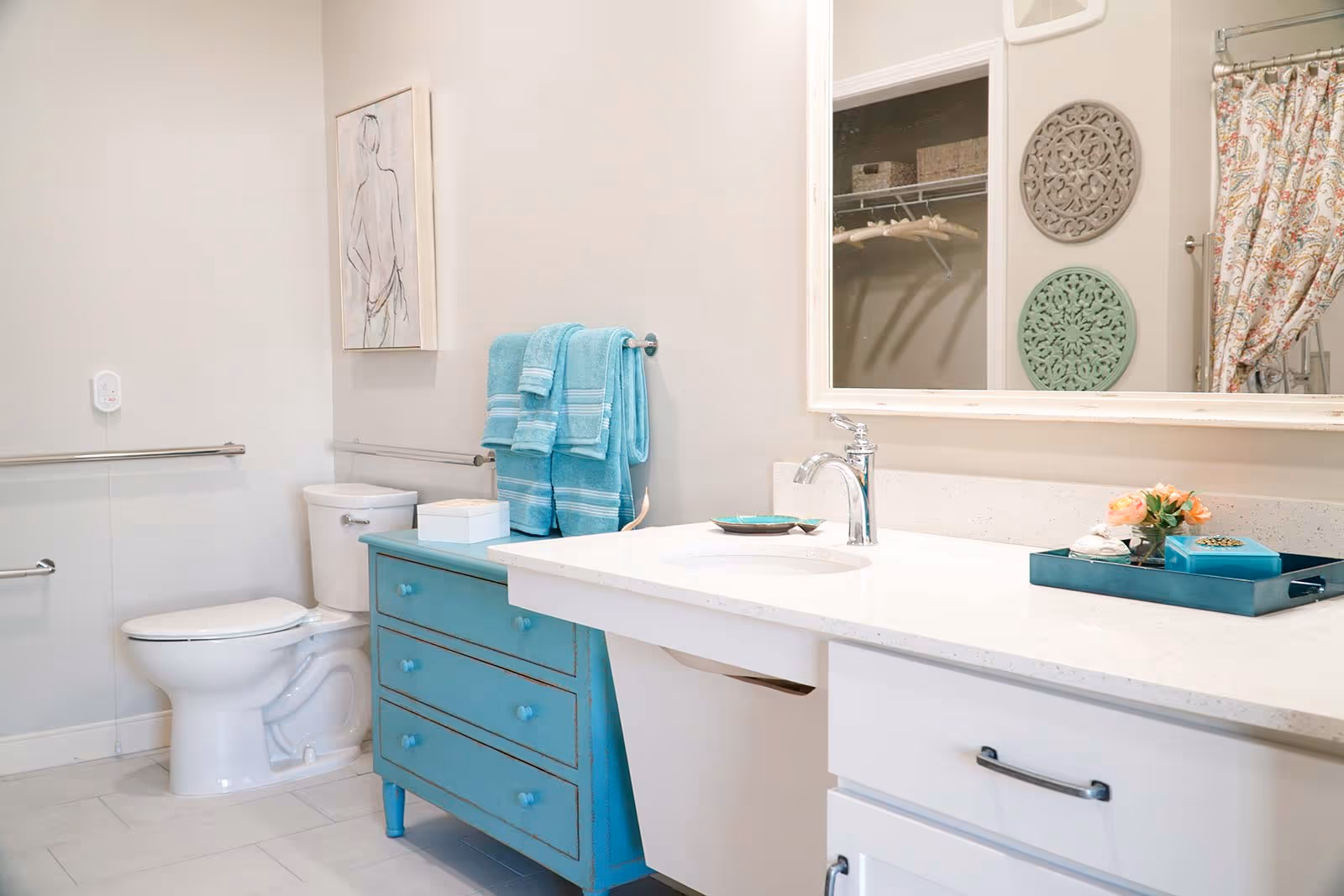 A bright bathroom featuring a white toilet, a turquoise blue dresser with matching towels, a white countertop with a sink and chrome faucet, a large mirror reflecting part of a closet and decorative wall hangings, and a floral shower curtain.