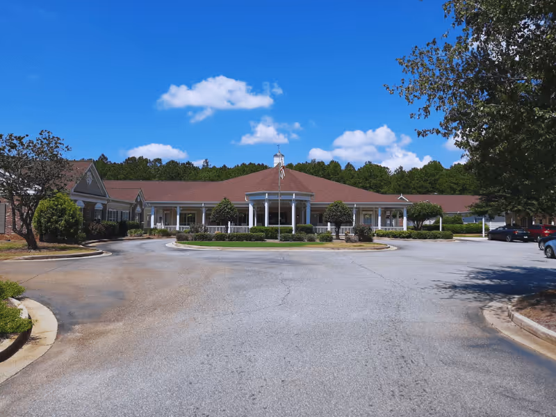 Front exterior view of a single-story senior living facility building with a red roof, white columns, and a circular driveway. There are trees and shrubs around the building and a clear blue sky with some clouds above.