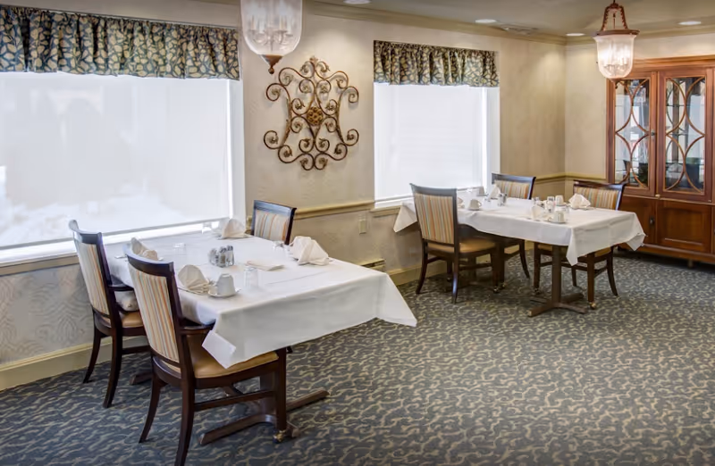 A dining room in a senior living facility with two tables covered in white tablecloths, each set with napkins, cups, and glasses. The room has patterned carpet, two large windows with patterned valances, a decorative wall piece, and a wooden china cabinet with glass doors. Two hanging light fixtures provide illumination.