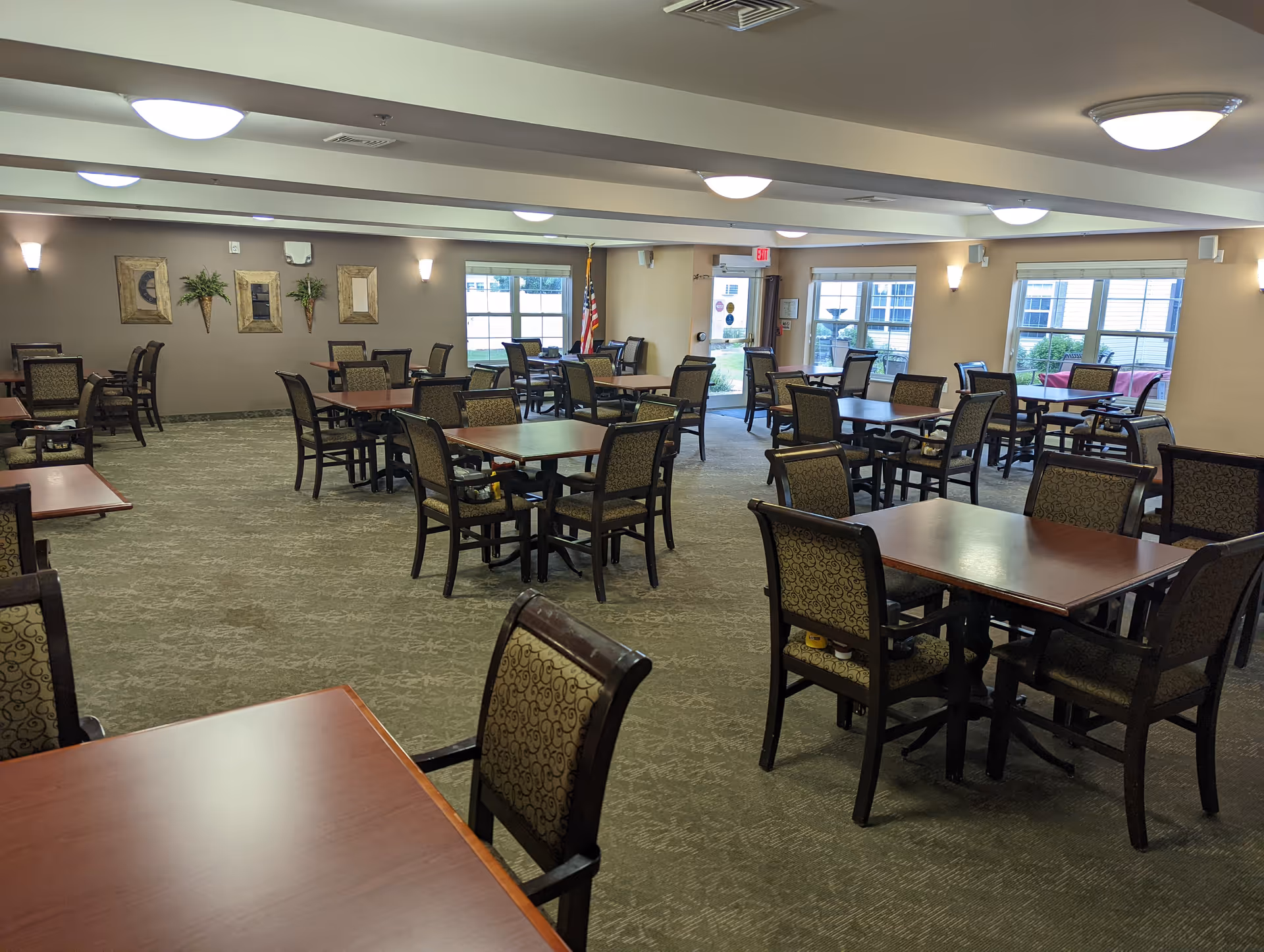 A spacious dining room with multiple square wooden tables and cushioned chairs arranged neatly. The room has large windows letting in natural light, beige walls decorated with framed art and plants, and ceiling lights providing additional illumination. An American flag is visible near the exit door.