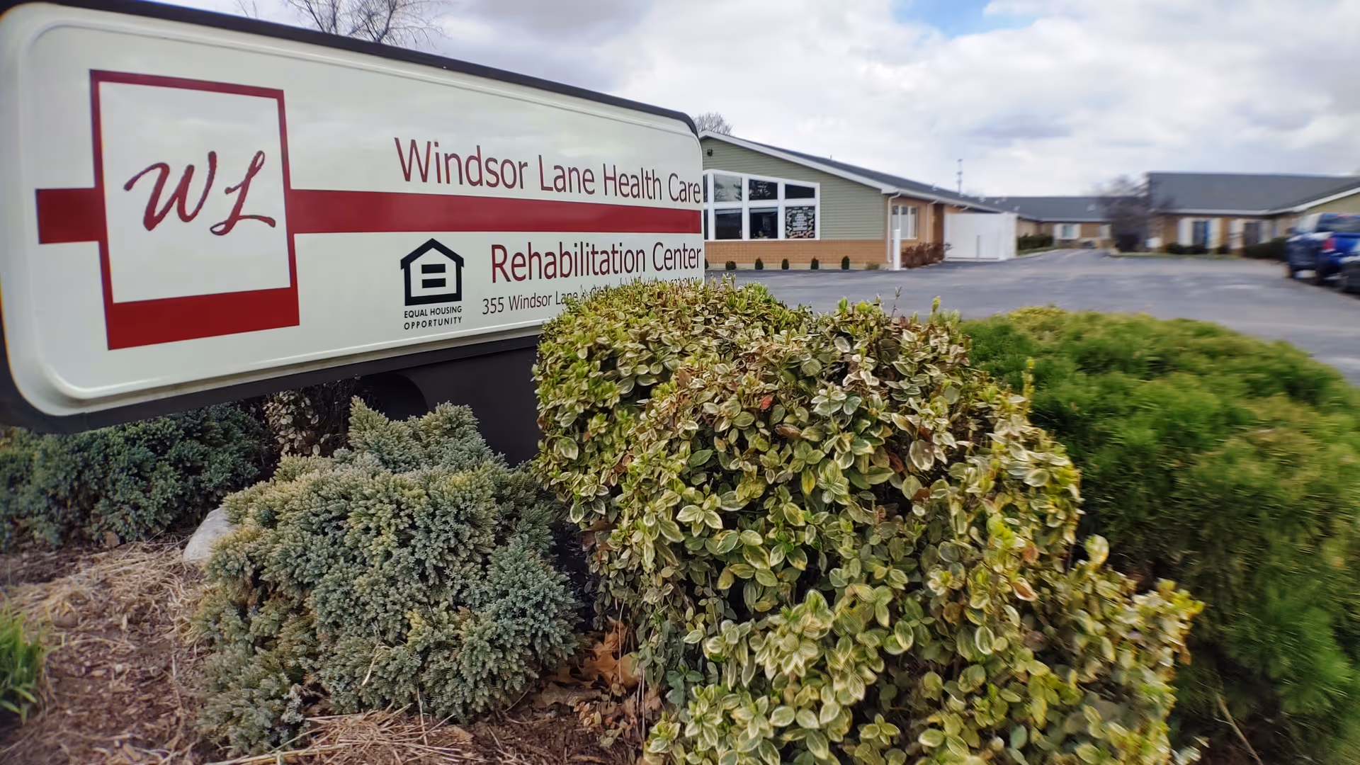 Outdoor view of Windsor Lane Health Care Rehabilitation Center sign surrounded by bushes and shrubs, with the building and parking lot visible in the background under a cloudy sky.