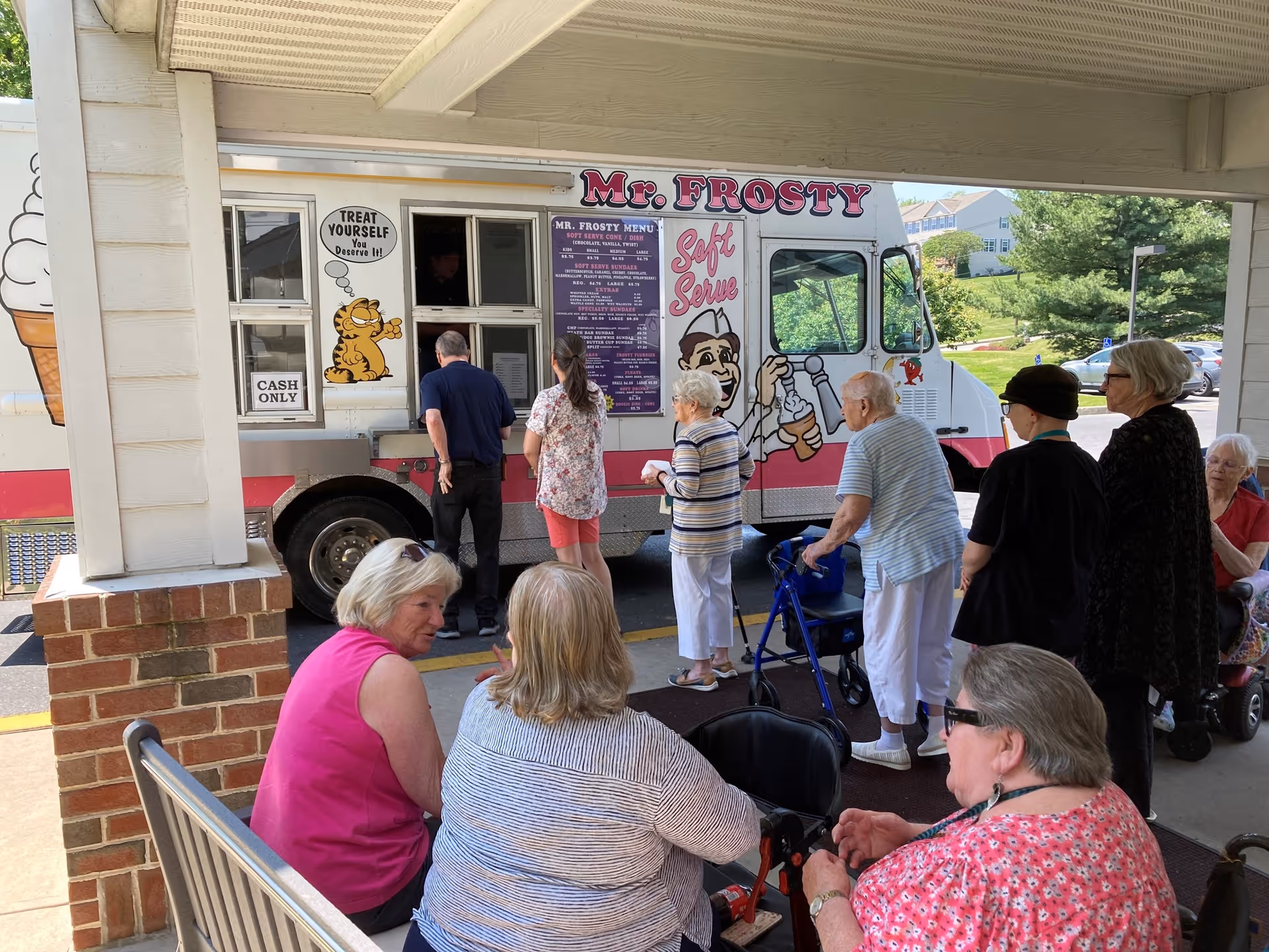 A group of elderly people and two younger adults are gathered outside near an ice cream truck named Mr. Frosty. Some are standing in line to order, while others are seated on benches under a covered area. The truck features colorful graphics including an ice cream cone and a cartoon character serving soft serve ice cream.