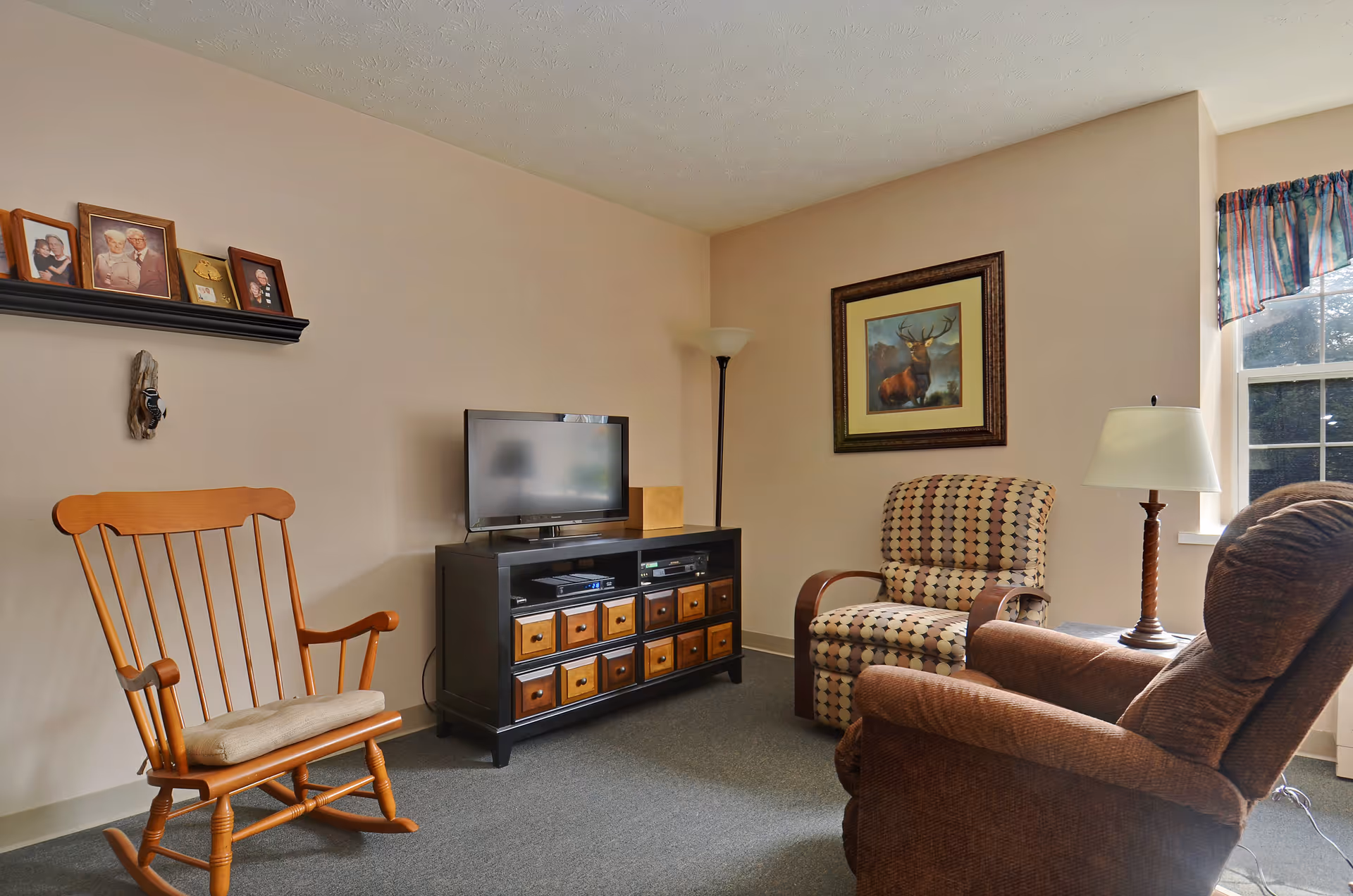 A cozy living room with a wooden rocking chair, a patterned armchair, and a brown recliner. There is a black TV stand with multiple drawers holding a flat-screen TV. A floor lamp stands in the corner, and a framed picture of a deer hangs on the wall. A window with a colorful valance lets in natural light, and a table lamp is placed on a small side table next to the recliner. A shelf on the wall holds framed photos and small decorative items.