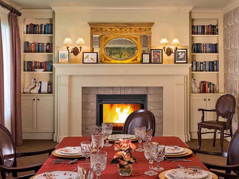 A cozy dining area with a table set for a meal, featuring floral-patterned plates, crystal glasses, and a small flower centerpiece. In the background, there is a lit fireplace with a decorative golden framed mirror above it, flanked by built-in bookshelves filled with books and decorative items. Two wall sconces with lamps provide warm lighting, and there are wooden chairs around the table and near the fireplace.