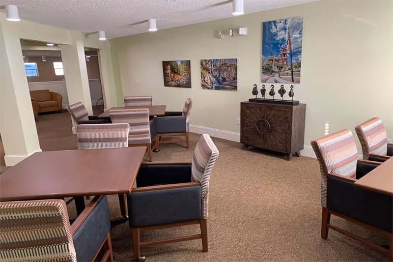 A well-lit common area with several wooden tables and cushioned chairs arranged around them. The walls are light-colored and decorated with three framed pictures and a dark wooden cabinet with decorative items on top. In the background, there is another seating area with a sofa.