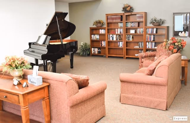 A cozy senior living common area featuring a grand piano, two upholstered sofas with floral cushions, wooden bookshelves filled with books, and floral arrangements on tables and shelves.