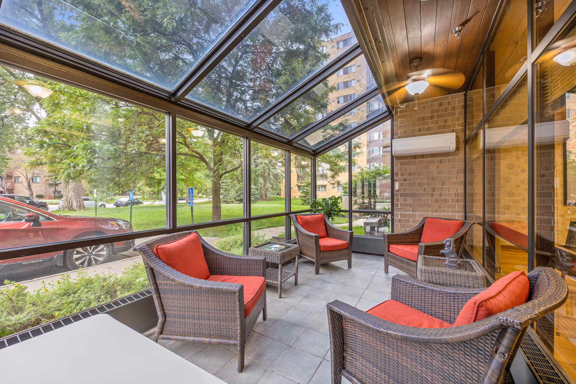 A sunroom with large glass windows and ceiling, furnished with four wicker chairs with red cushions arranged around two small glass-top tables. The room has a tiled floor, a ceiling fan, and views of green trees and a red car outside.