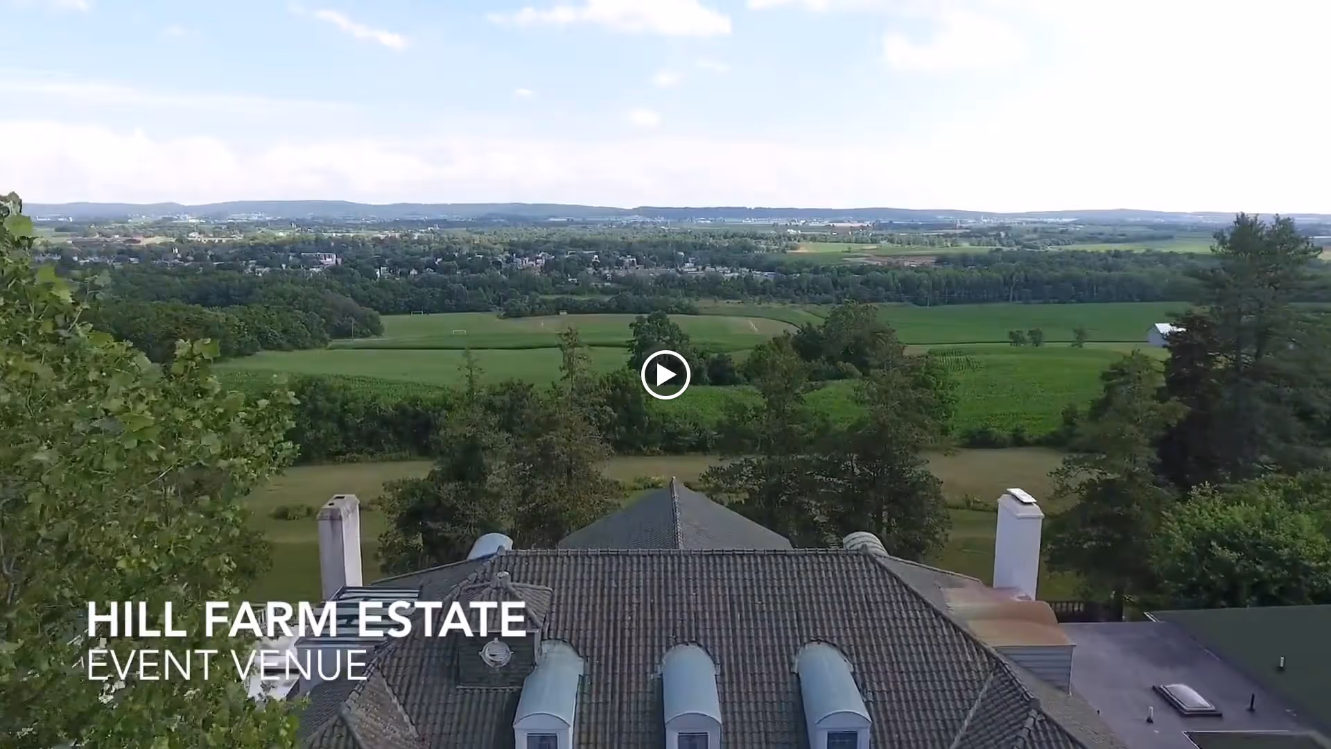 Aerial view of a large building with a tiled roof surrounded by trees and expansive green fields extending into the distance under a partly cloudy sky. Text on the image reads 'Hill Farm Estate Event Venue'.