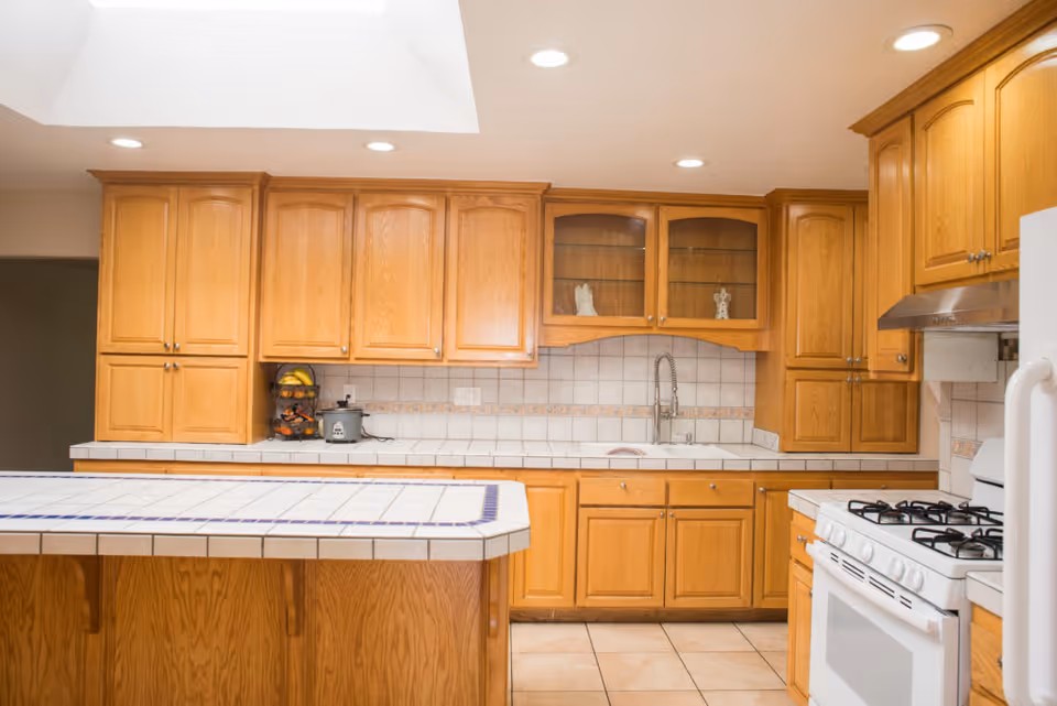 A bright kitchen with wooden cabinets, a tiled countertop island, a white gas stove with oven, and a double sink under a tiled backsplash. The kitchen has recessed lighting and a skylight above.