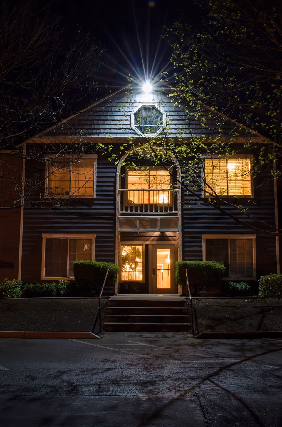 Night view of the front of a two-story residential building with illuminated windows, a central doorway, and an overhead exterior light.