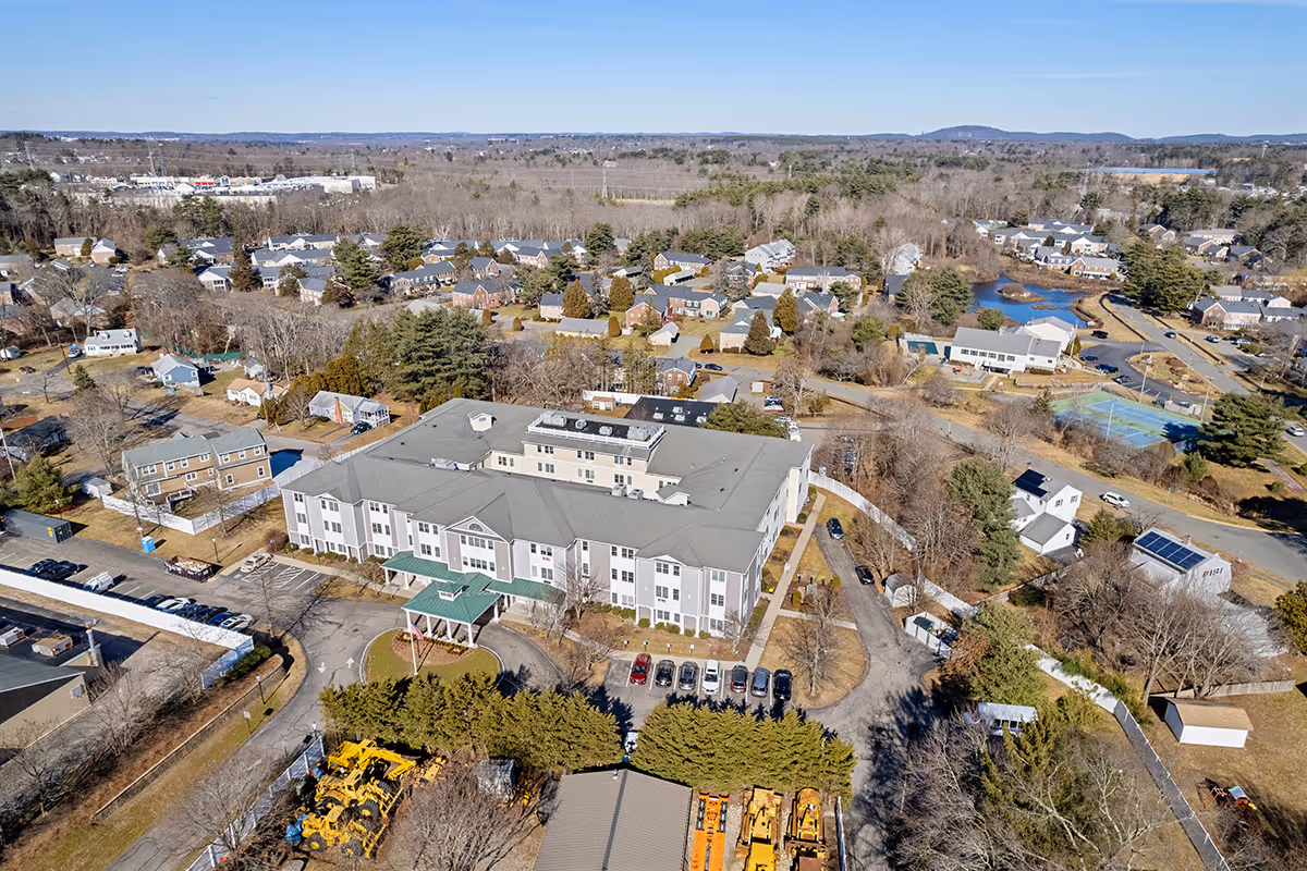 Aerial view of a large senior living facility building with a green entrance canopy, surrounded by parking lots, trees, and residential houses in the background under a clear sky.