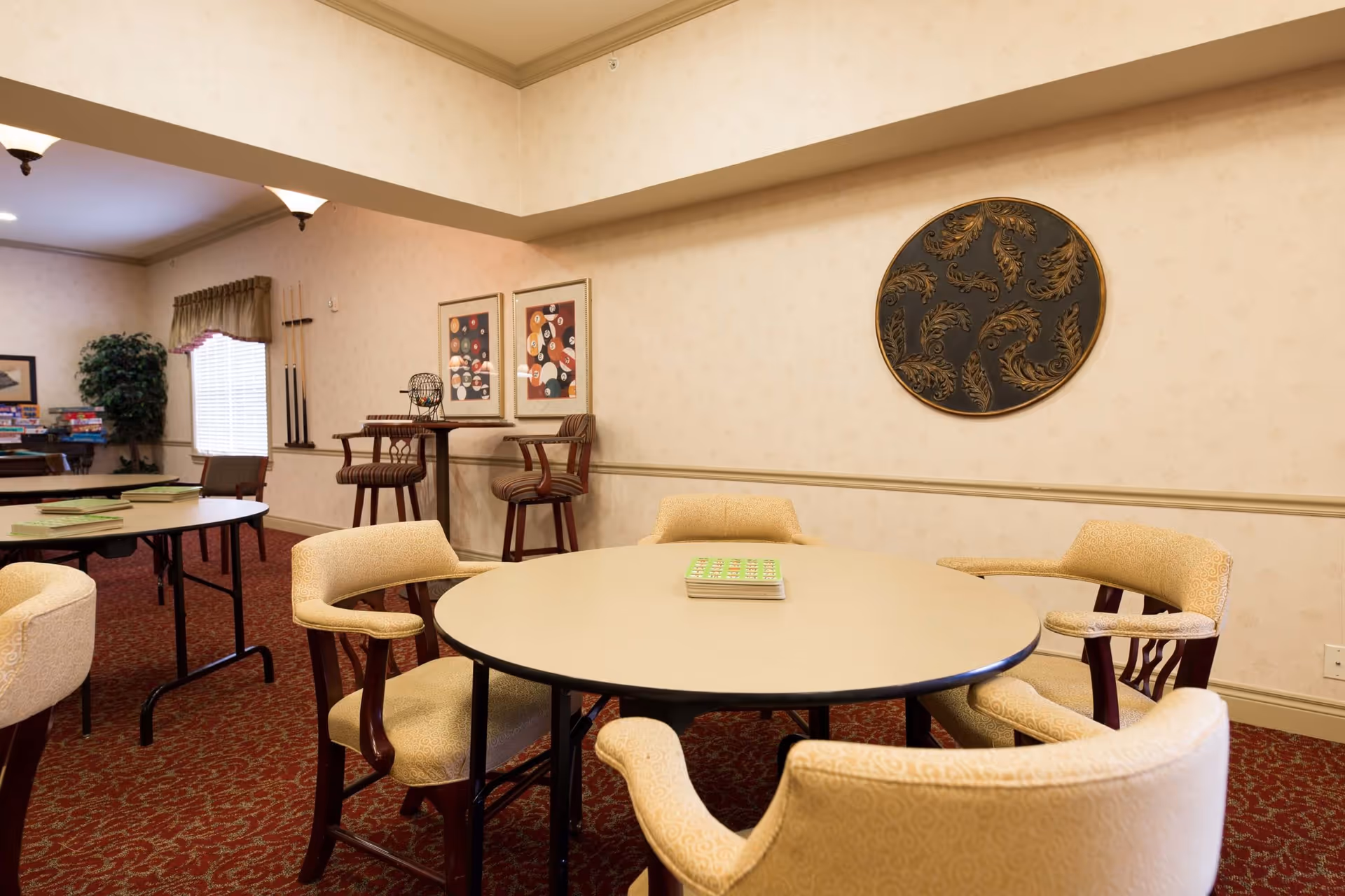 A cozy activity room with a round table surrounded by six beige upholstered chairs. On the table, there is a bingo card. The room has red patterned carpet, beige walls with decorative molding, and framed artwork on the walls. In the background, there are additional tables, chairs, a window with curtains, a potted plant, and a rack holding pool cues.