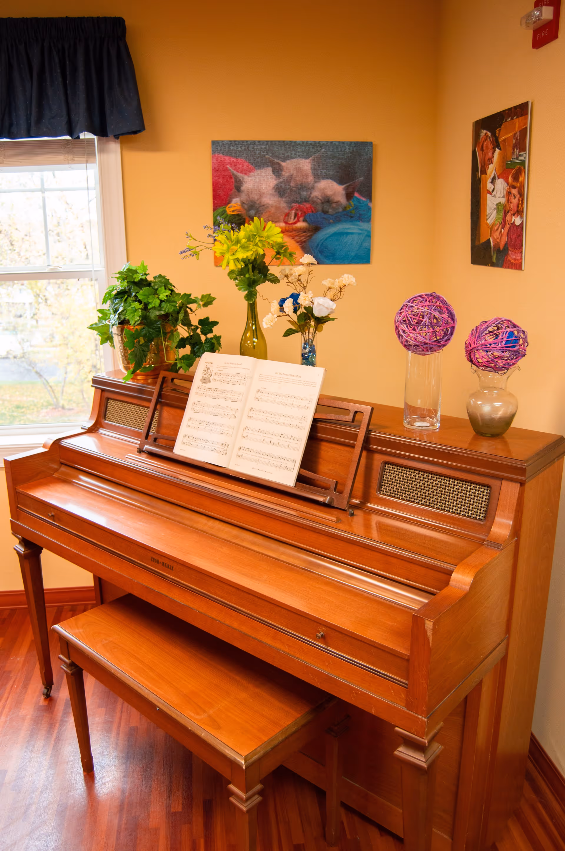 A wooden upright piano with an open sheet of music on the stand, a matching wooden bench, and decorative items on top including two vases with flowers and two purple woven balls in glass containers. The piano is placed against a beige wall with two framed pictures hanging above it, and a window with a dark curtain is visible to the left.