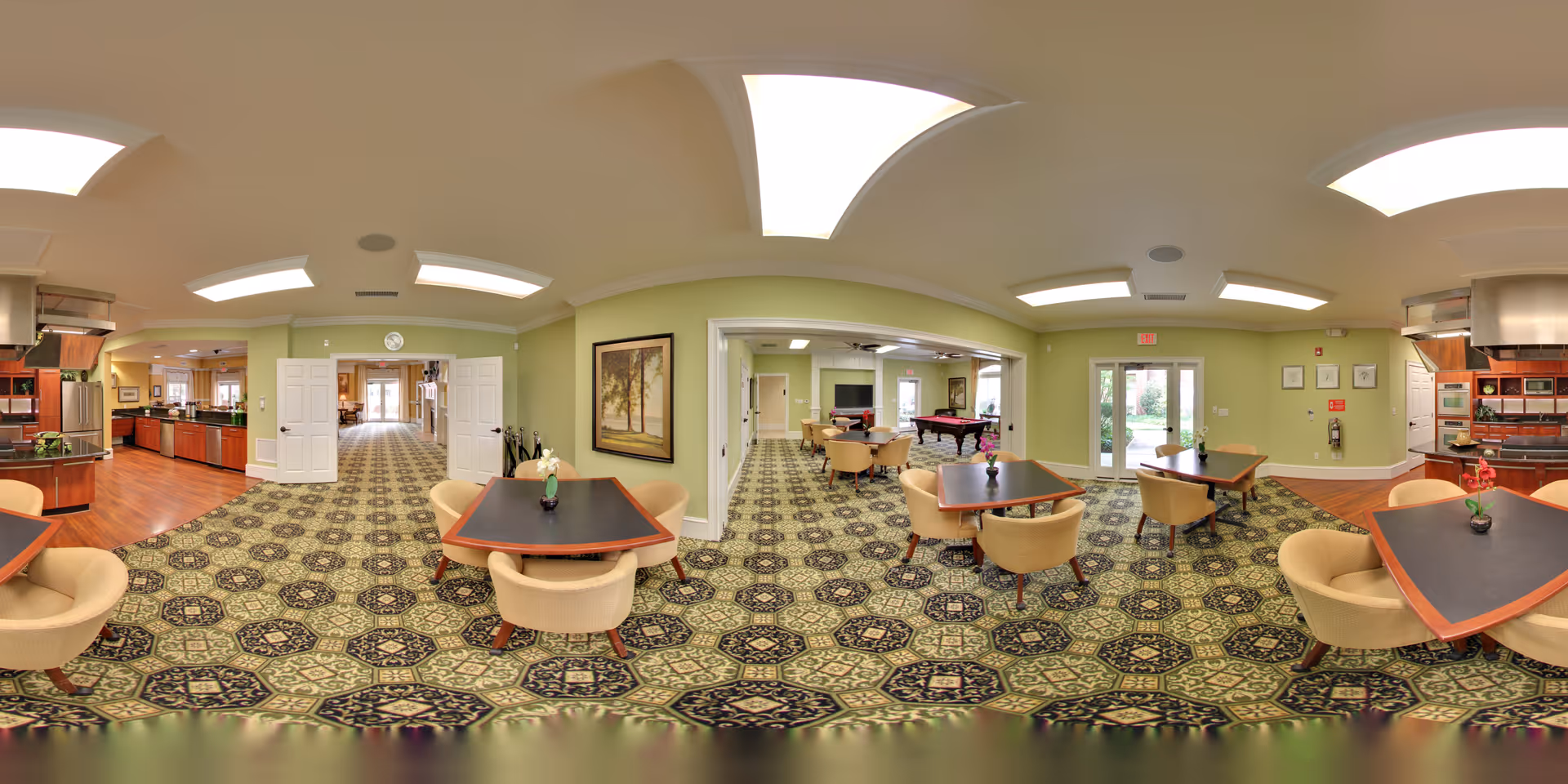 A spacious and well-lit common area in a senior living facility featuring multiple tables with beige chairs arranged around them. The room has patterned carpet flooring and light green walls. There is a kitchen area with stainless steel appliances and wooden cabinetry on the left side. In the background, there is a room with a pool table and additional seating. Large windows and glass doors provide natural light and a view of the outdoor greenery.
