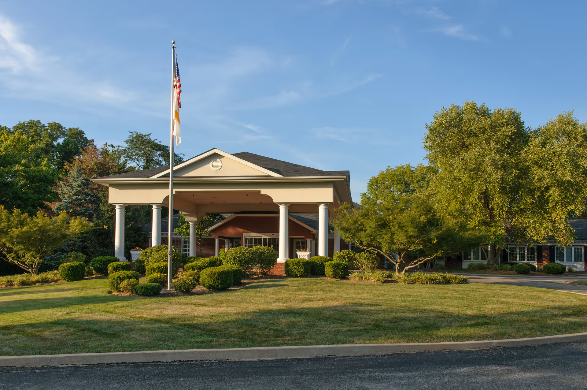 Entrance of The Westchester House showing a columned portico, flagpole, manicured lawn, and surrounding trees.