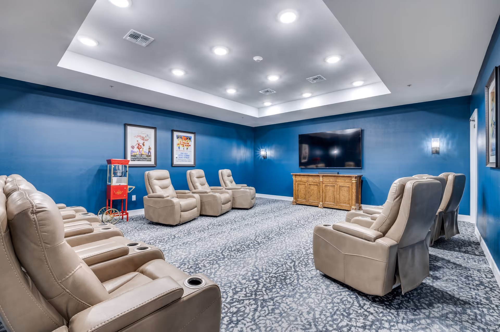 A cozy media room with beige recliner chairs arranged in two rows facing a large flat-screen TV mounted on a wooden cabinet. The walls are painted blue and decorated with framed posters. A popcorn machine is placed against one wall. The ceiling has recessed lighting and a tray design.