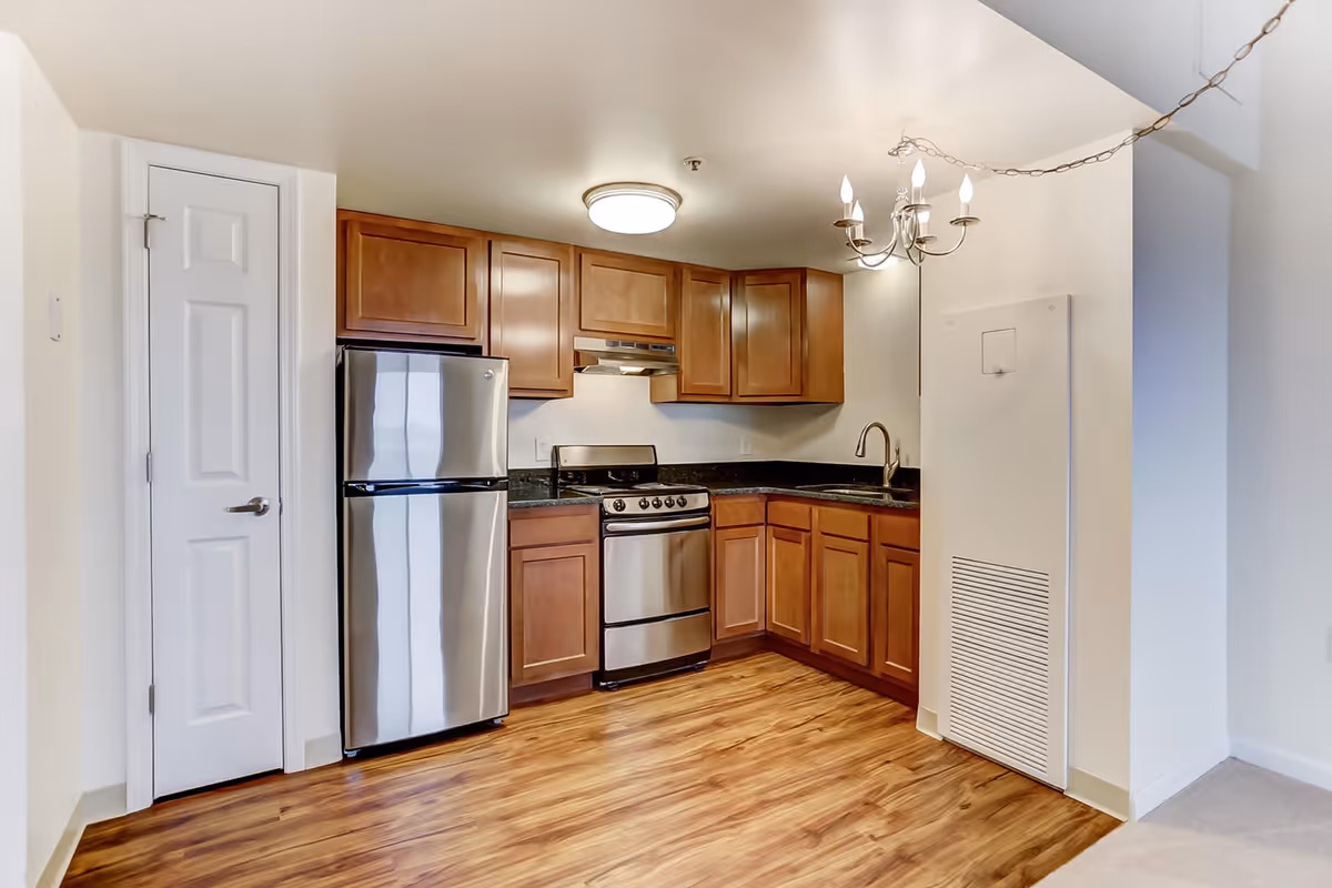 A modern kitchen with wooden cabinets, a stainless steel refrigerator, stove, and sink. The floor is wood laminate, and there is a white door to the left and a chandelier light fixture hanging from the ceiling.