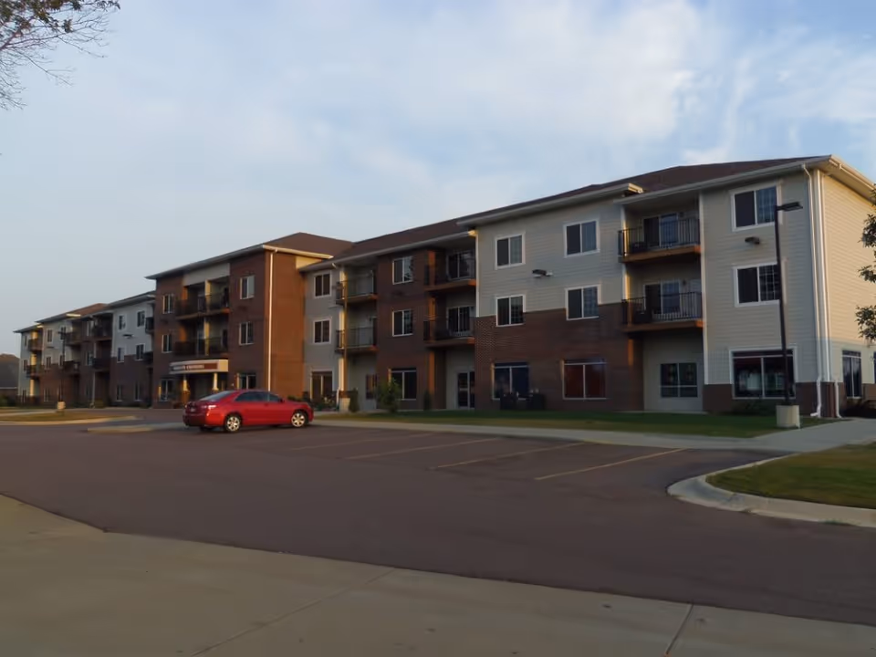 Three-story apartment-style senior living building with balconies, a parking lot and a red car in front under a blue sky.