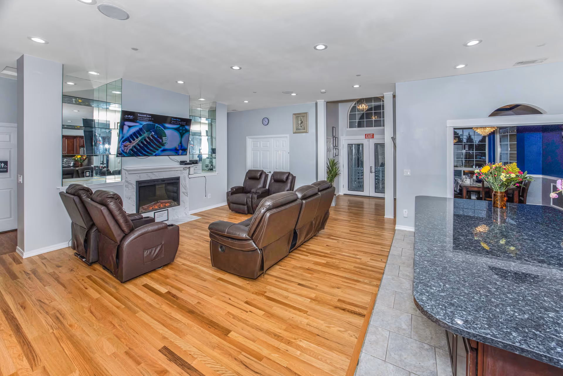 Spacious common living area with leather recliners arranged around a fireplace and wall-mounted TV, hardwood floors, and a granite countertop in the foreground.