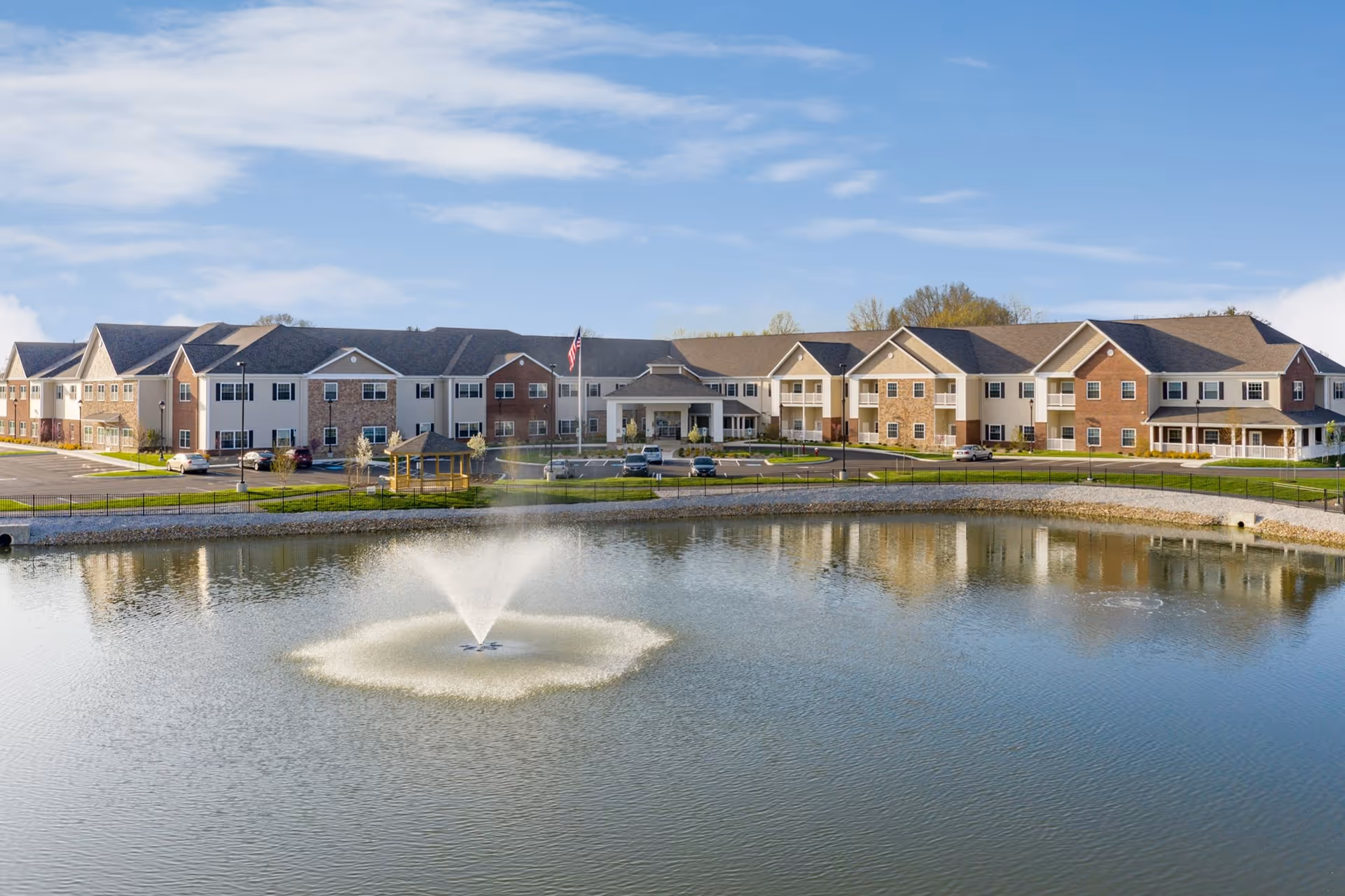 Exterior view of a large senior living building with a central entrance, parking lot, and a pond with a fountain in the foreground.