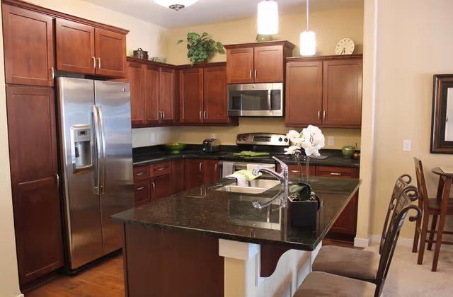 Modern kitchen with dark granite countertops, wooden cabinets, stainless steel refrigerator, microwave, and stove. There is a kitchen island with a sink and two chairs, and decorative plants on top of the cabinets and island.