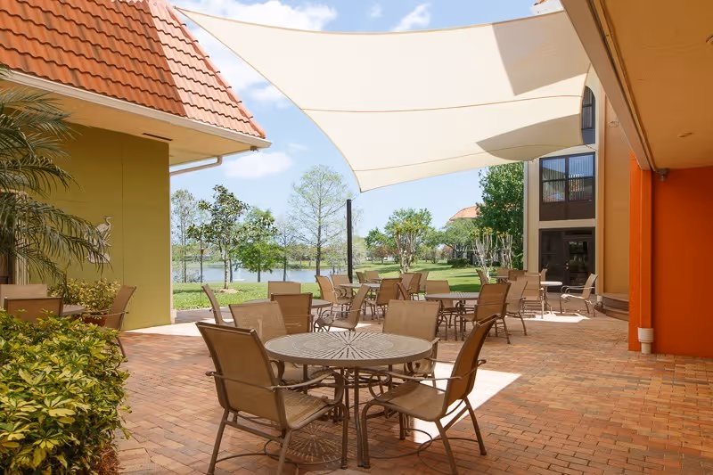 Outdoor patio area with multiple round tables and chairs under a large white shade sail. The patio is paved with bricks and surrounded by greenery, trees, and a view of a lake in the background. The buildings around the patio have orange and green walls with tiled roofs.