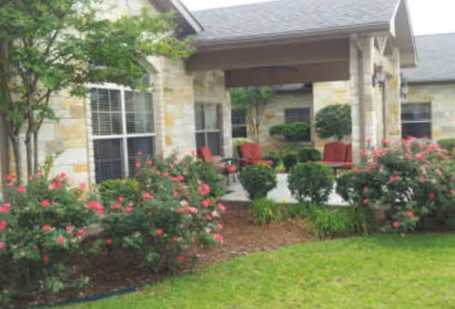 Outdoor patio area of a stone building with large windows, surrounded by green bushes and pink flowering plants. The patio has several red cushioned chairs arranged around a small table under a covered roof.