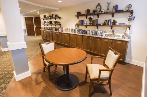 A cozy interior space with a round wooden table and two cushioned chairs on a wooden floor. Behind the table is a long wooden cabinet topped with a marble surface, decorated with books, vases, and ornamental items. Above the cabinet are two wooden shelves holding more books and decorative pieces. The area is well-lit with recessed ceiling lights and has a carpeted hallway leading to closed doors.