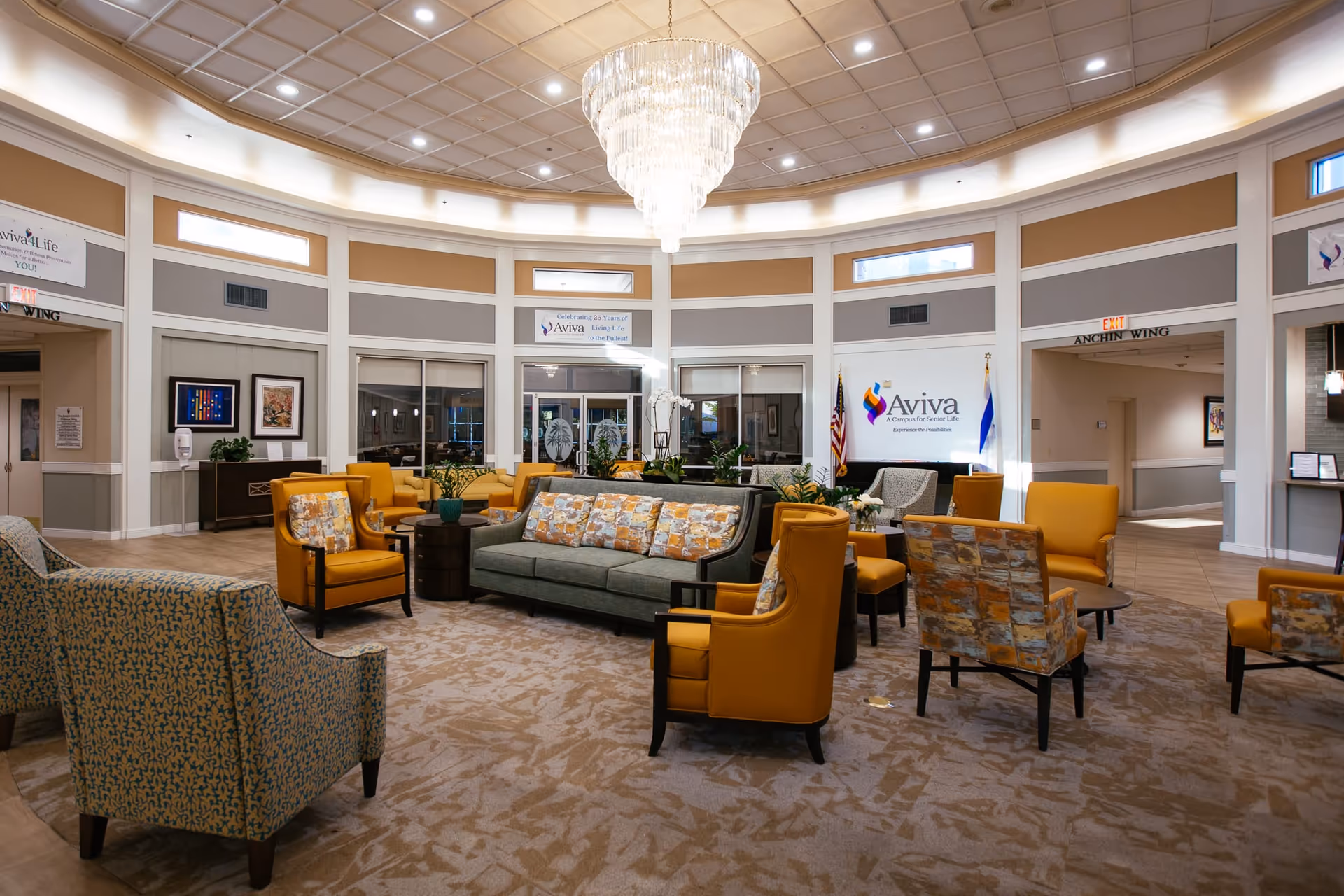 A spacious and well-lit senior living facility common area with a large chandelier hanging from a high ceiling. The room features multiple comfortable chairs and sofas in yellow and patterned upholstery arranged around small tables on a carpeted floor. The walls have windows near the ceiling and signs with the Aviva Senior Living logo and slogans. There are American and other flags near the wall, and doorways leading to other parts of the facility.