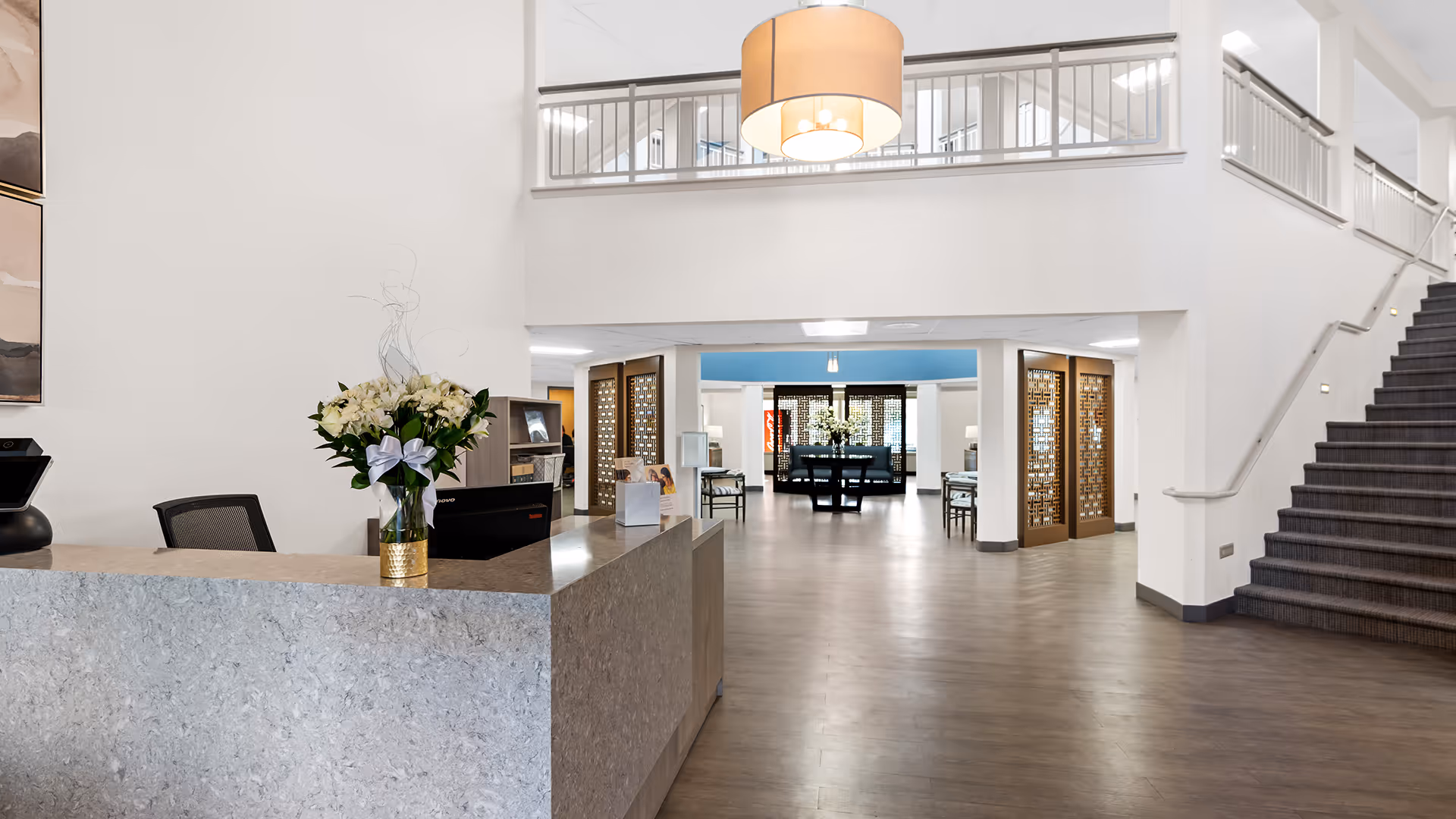 A spacious and modern reception area with a large stone front desk adorned with a vase of white flowers. Behind the desk is a chair and office equipment. The area features high ceilings with a large hanging light fixture, a staircase with carpeted steps on the right, and an open hallway leading to a seating area with tables and chairs.