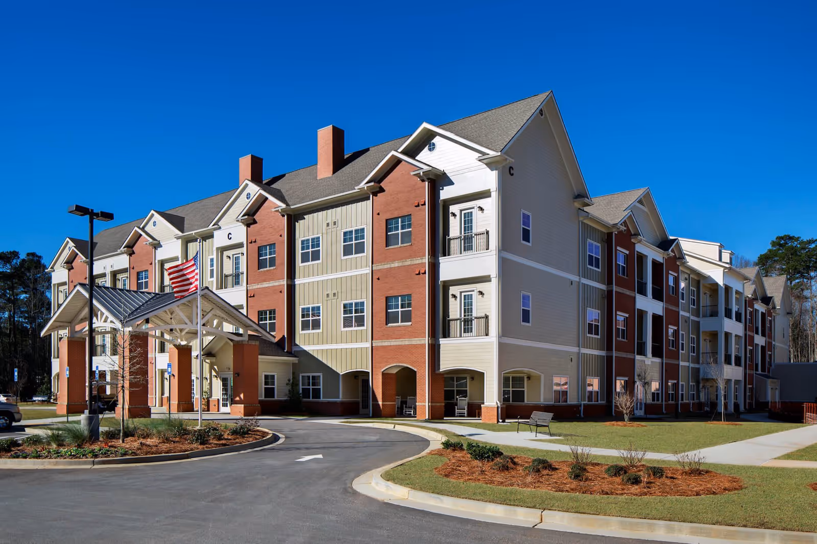 Exterior view of a multi-story senior living facility building with a covered entrance, an American flag, and landscaped grounds under a clear blue sky.